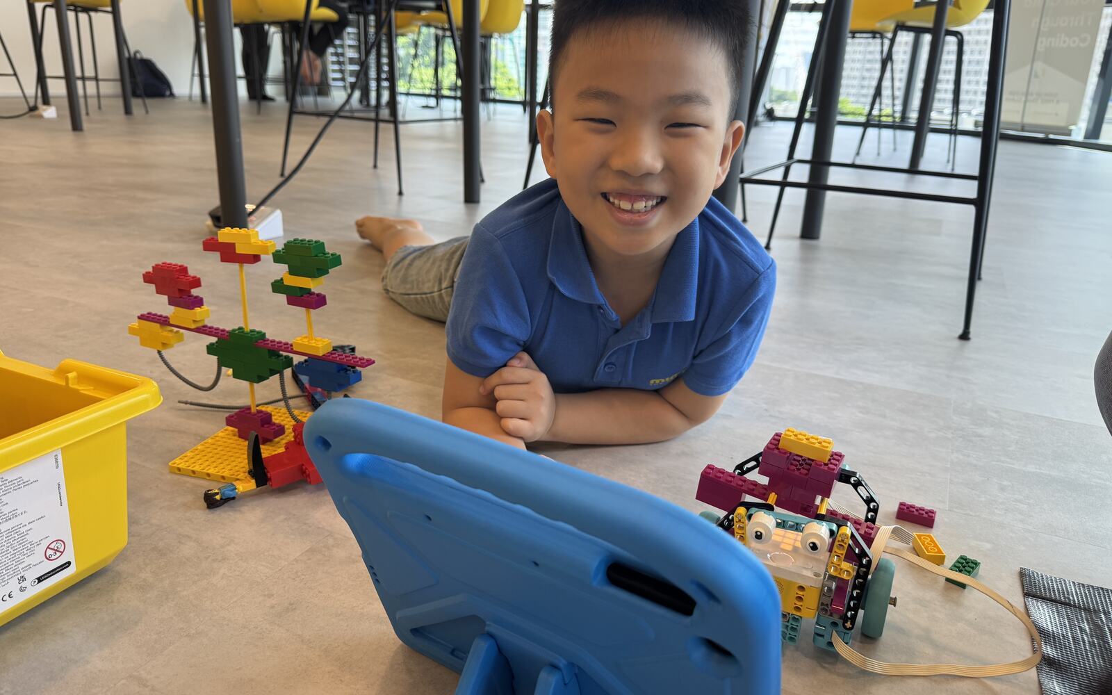 A Lab student smiling on the floor beside his finished LEGO robotics build and a coding tablet